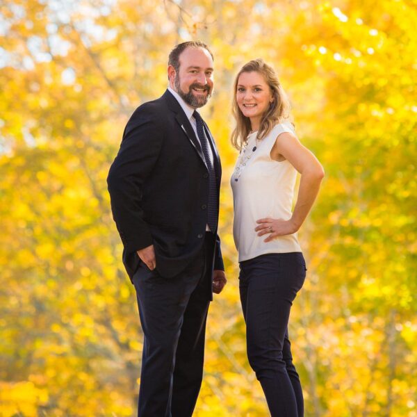 A man and a woman stand side by side in a vibrant outdoor setting filled with golden autumn leaves. The man is dressed in a black suit with a tie, sporting a beard and a friendly smile. The woman wears a light-colored top with decorative beads and dark pants, also smiling confidently. They both pose with relaxed postures, exuding a sense of warmth and professionalism against the colorful backdrop.