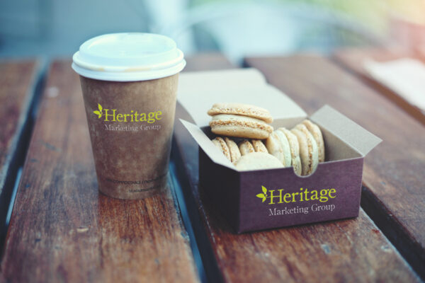 A disposable coffee cup with a white lid features a logo for "Heritage Marketing Group." Next to it is a small brown box containing several macarons in various shades, including beige and light green. The items are placed on a wooden surface, suggesting a casual setting.