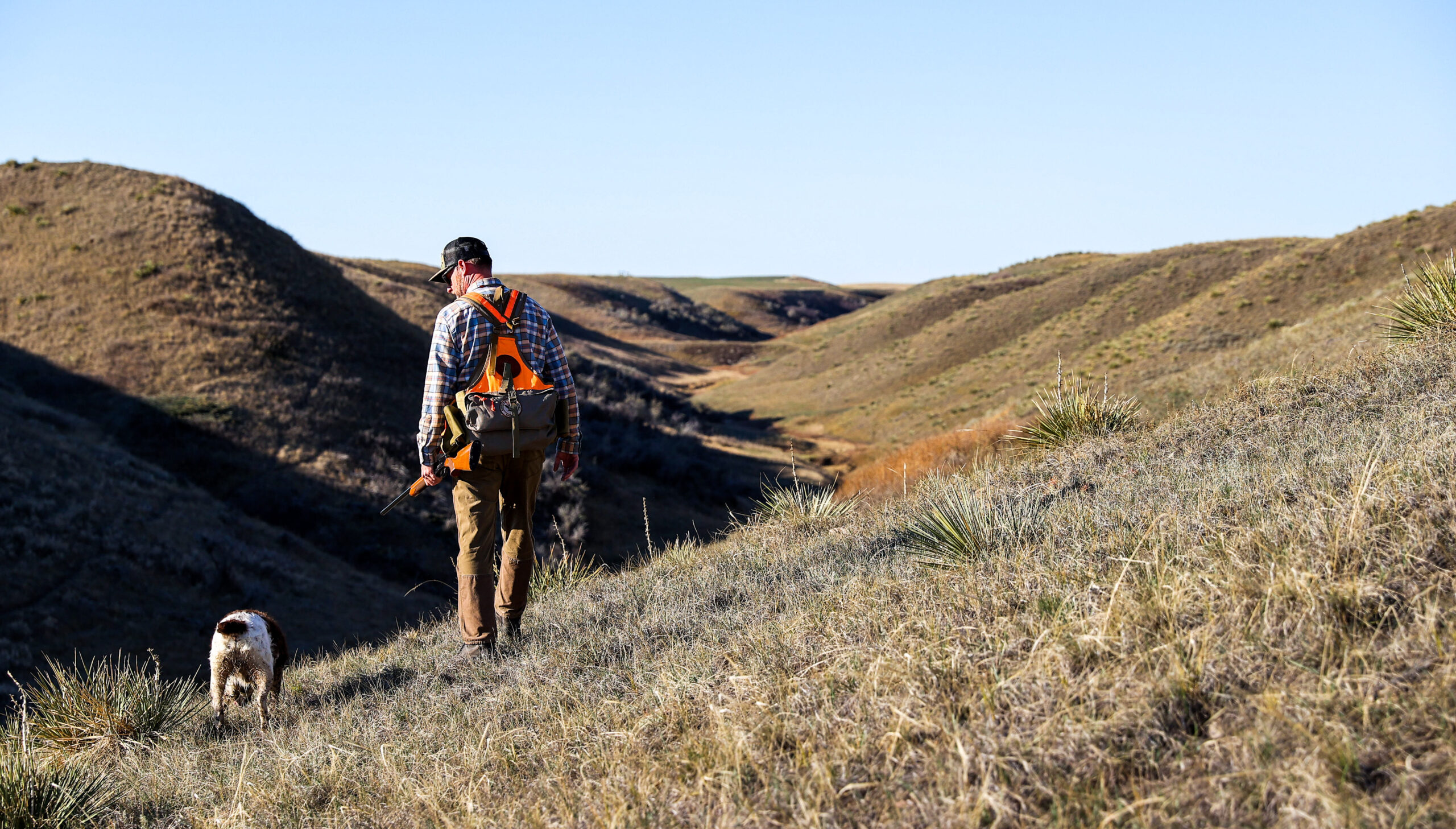 Young man with hunting gear walking on the side of a hill with his dog