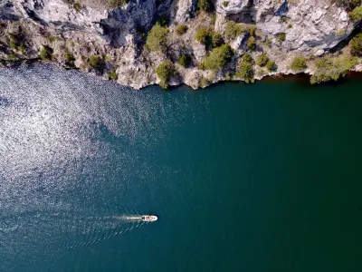 Aerial view of a tranquil body of water bordered by rocky cliffs. The water is a deep blue with shimmering reflections from sunlight. A small boat is seen moving across the water, leaving a gentle wake behind it. The cliffs are dotted with patches of greenery, adding contrast to the rugged landscape.