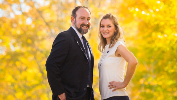 A man and a woman are posing together outdoors against a backdrop of vibrant autumn foliage. The man is dressed in a black suit with a white shirt and a patterned tie, smiling confidently. The woman is wearing a white top with a necklace and dark pants, also smiling and standing with her hand on her hip. The warm colors of the leaves create a cheerful atmosphere.