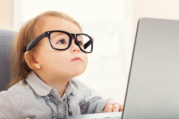 A young child with light brown hair is sitting in front of a laptop, wearing large black glasses. The child is focused, gazing upwards with a thoughtful expression. They are dressed in a striped shirt with a collar, and their hand is resting on the laptop keyboard. The background is softly blurred, suggesting a bright and airy indoor environment.
